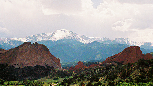 [Image includes three layers of rock. In the far distance are the highest peaks which are covered in snow. In the middle distance are shorter mountains which are covered in greenery. In the foreground are the red rocks which only have vegetation at the foot of them. There is a low section of red rock in the middle of the image creating an opening to more clearly see the middle distance mountains. The sky is nearly completely covered in white clouds although the foreground of the image is very sunny.]